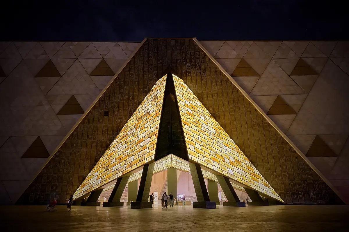 The glowing alabaster-clad entrance of the Grand Egyptian Museum under a wall of hieroglyphic cartouches at night